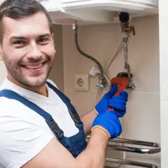 Uśmiechnięty hydraulik w niebieskim kombinezonie naprawia bojler, który nie grzeje wody. Używa klucza nastawnego.