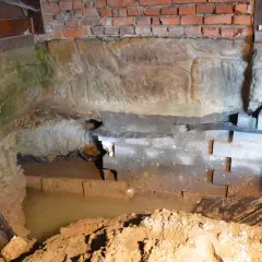 W piwnicy wybija woda, zalewając fundamenty. Widać wilgoć na cegłach i kamieniach.