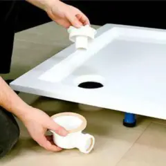 Montaż brodzika z konglomeratu na płytkach. Ręce instalatora montują syfon pod brodzikiem.