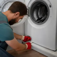Mężczyzna w czerwonych rękawiczkach próbuje otworzyć pralkę awaryjnie, odkręcając filtr.