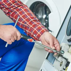 Technik w niebieskim kombinezonie i kratowanej koszuli naprawia pralkę, używając klucza nasadowego.
