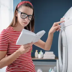 Kobieta z okularami i czerwoną opaską na włosach czyta instrukcję, próbując rozwiązać problem z pralką, być może błąd E10.