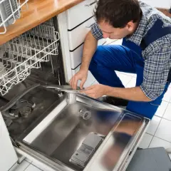 Technik w niebieskim kombinezonie pochyla się nad otwartą zmywarką, pokazując, jak podłączyć zmywarkę. Obok leżą klucze i rękawice.