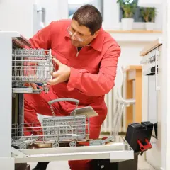 Serwisant w czerwonym kombinezonie naprawia zmywarkę, która ma zatkany odpływ. W rękach trzyma narzędzia.