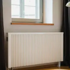 Biały grzejnik pod oknem, obok ciemnej zasłony i lampy stojącej. Dowiedz się, jak ustawić grzejniki przy ogrzewaniu gazowym dla optymalnego komfortu.