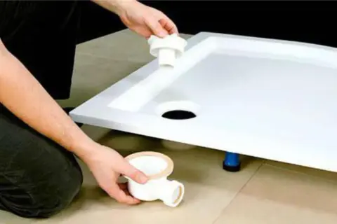 Montaż brodzika z konglomeratu na płytkach. Ręce instalatora montują syfon pod brodzikiem.