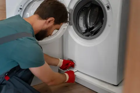 Mężczyzna w czerwonych rękawiczkach próbuje otworzyć pralkę awaryjnie, odkręcając filtr.