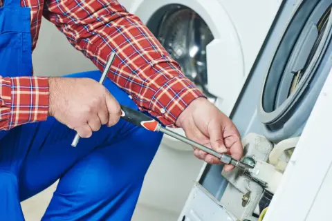 Technik w niebieskim kombinezonie i kratowanej koszuli naprawia pralkę, używając klucza nasadowego.