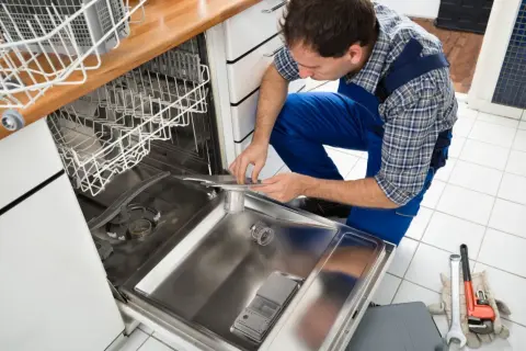 Technik w niebieskim kombinezonie pochyla się nad otwartą zmywarką, pokazując, jak podłączyć zmywarkę. Obok leżą klucze i rękawice.
