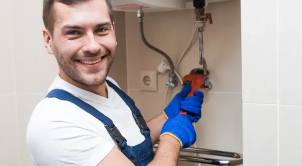 Uśmiechnięty hydraulik w niebieskim kombinezonie naprawia bojler, który nie grzeje wody. Używa klucza nastawnego.