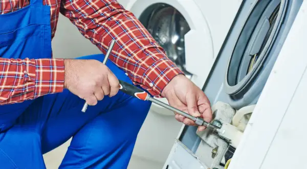 Technik w niebieskim kombinezonie i kratowanej koszuli naprawia pralkę, używając klucza nasadowego.