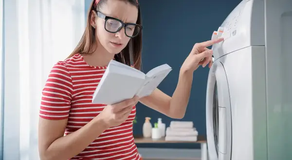 Kobieta z okularami i czerwoną opaską na włosach czyta instrukcję, próbując rozwiązać problem z pralką, być może błąd E10.