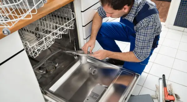 Technik w niebieskim kombinezonie pochyla się nad otwartą zmywarką, pokazując, jak podłączyć zmywarkę. Obok leżą klucze i rękawice.