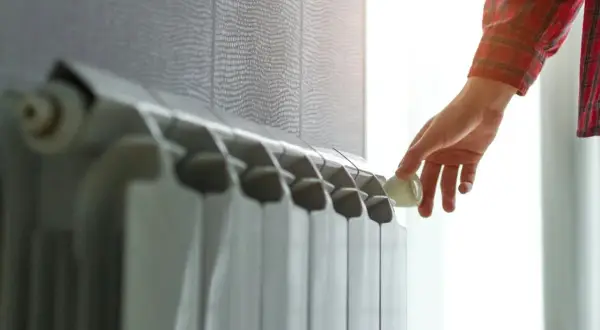 Dłoń reguluje termostat na grzejniku aluminiowym, który współpracuje z pompą ciepła, zapewniając komfort cieplny.
