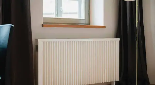 Biały grzejnik pod oknem, obok ciemnej zasłony i lampy stojącej. Dowiedz się, jak ustawić grzejniki przy ogrzewaniu gazowym dla optymalnego komfortu.