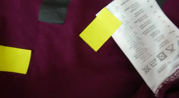 Małe dziury w ubraniach po praniu, widoczne na bordowym materiale, obok żółtych karteczek i metki.
