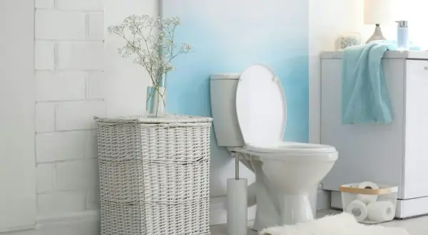Łazienka z białym, ceramicznym sedesem i otwartą klapą. Obok kosz z wikliny, a za nim spłuczka starego typu.