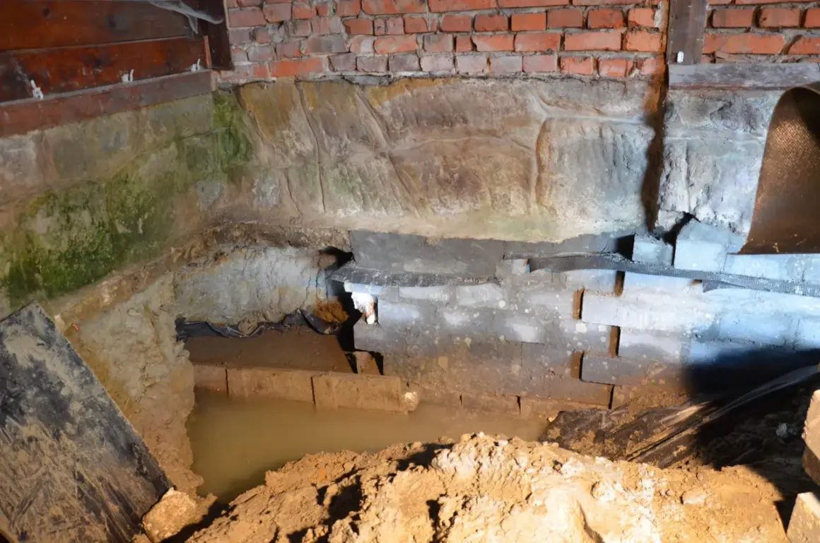 W piwnicy wybija woda, zalewając fundamenty. Widać wilgoć na cegłach i kamieniach.