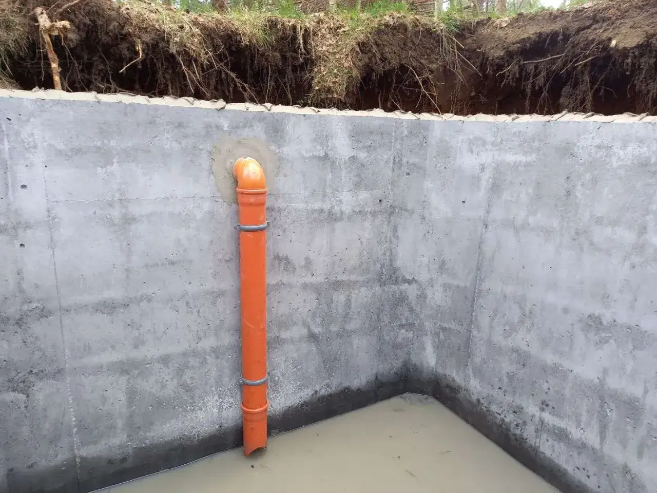 Betonowy zbiornik, gotowy na pozwolenie na szambo. Pomarańczowa rura odpływowa zanurzona w wodzie.