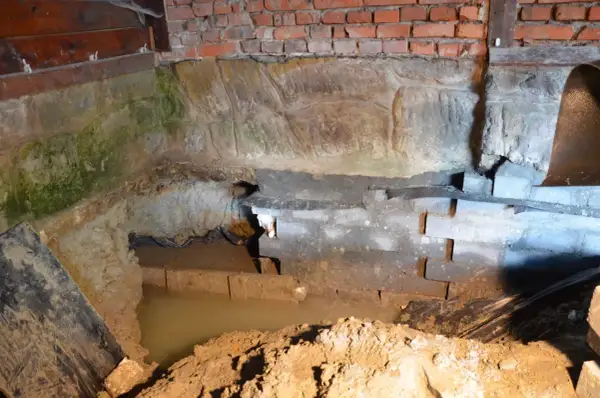 W piwnicy wybija woda, zalewając fundamenty. Widać wilgoć na cegłach i kamieniach.