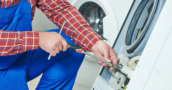 Technik w niebieskim kombinezonie i kratowanej koszuli naprawia pralkę, używając klucza nasadowego.
