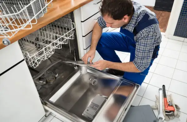 Technik w niebieskim kombinezonie pochyla się nad otwartą zmywarką, pokazując, jak podłączyć zmywarkę. Obok leżą klucze i rękawice.