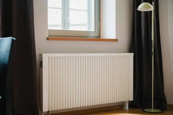 Biały grzejnik pod oknem, obok ciemnej zasłony i lampy stojącej. Dowiedz się, jak ustawić grzejniki przy ogrzewaniu gazowym dla optymalnego komfortu.